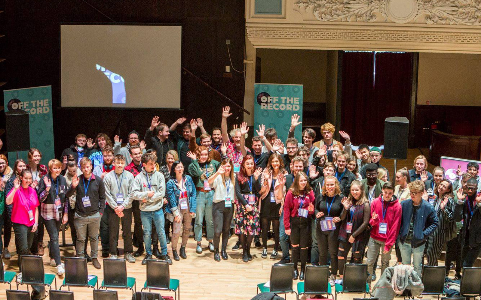 A large group of young people crowded together on a stage. Pop-up banners surrounding them read Off The Record.