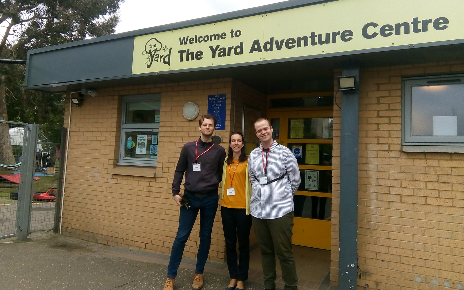 Three people standing together at the front entrance of a building. Above the door a sign reads Welcome to The Yard Adventure Centre