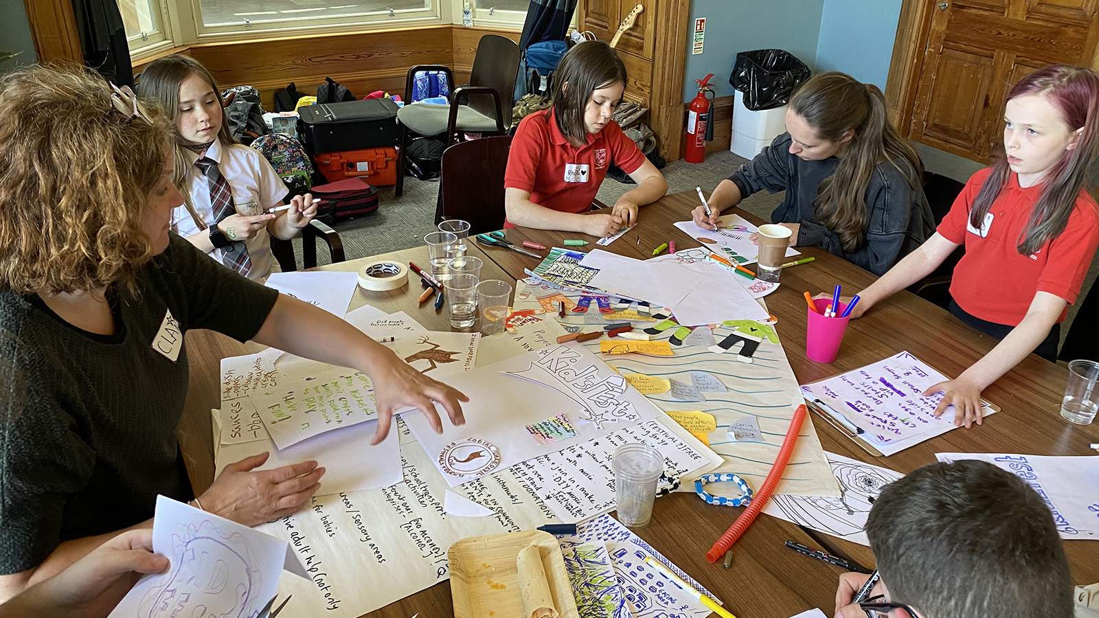 A group of children sitting around a large table covered in paper and pens, some writing, some drawing. They are talking to a woman who is showing them how to create a collage.