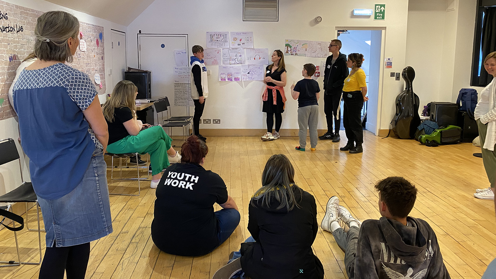 A group of young people and adults in a large well lit room with large pieces of paper on the wall covered in writing. One young person standing next to drawings on the wall is speaking, everyone else is watching and listening.