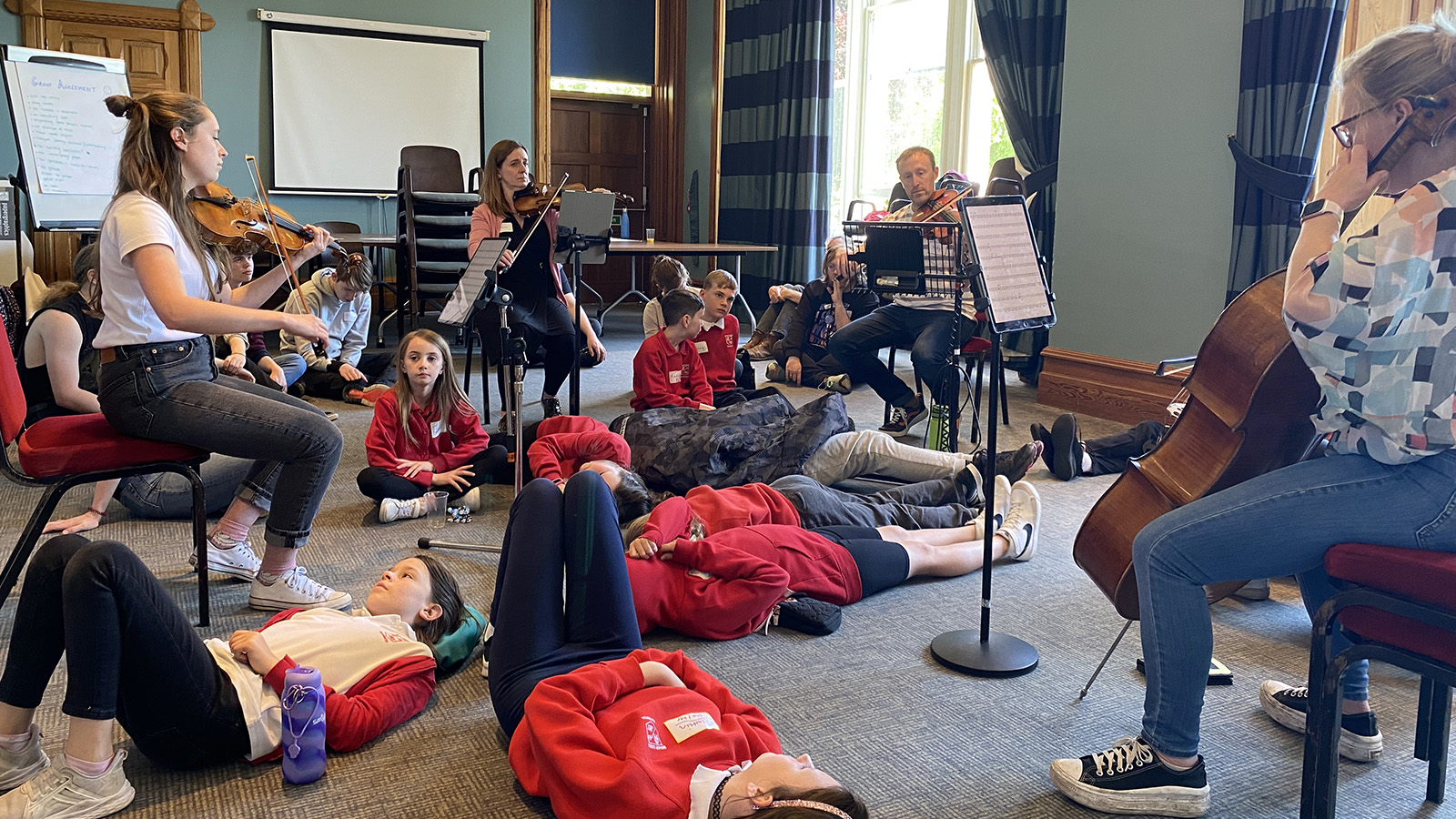 Group of children in school uniform sitting and lying on the floor while listening and watching a live performance of musicians playing violins.