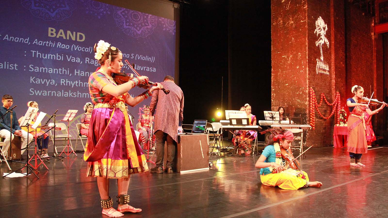 Three young musicians playing violins and wearing traditional Indian dress . Two are standing on the right and left and one is seated. Behind them a band sits next to their instruments - they are listening to the other musicians. A large projection behind them names the musicians.