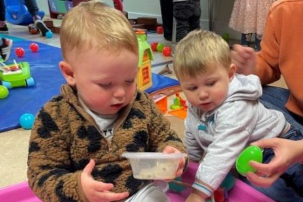 Two young boys in a colourful, busy playroom play with a 'shaker' box while an adult holds a bright green coloured egg shaker