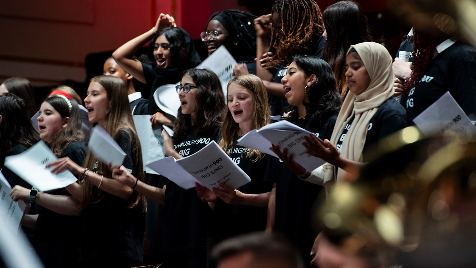A diverse choir performs together, wearing matching black shirts and holding music sheets, in a vibrant atmosphere.