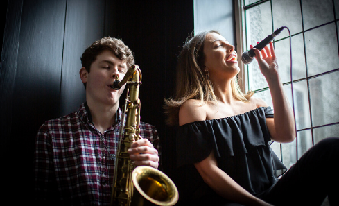 A man plays saxophone and a woman sings into a microphone as they sit in the light of a window together
