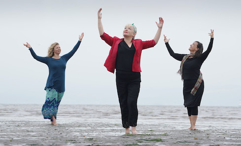 Three dancers stand with their arms outstretched overhead on a grey day on a beach