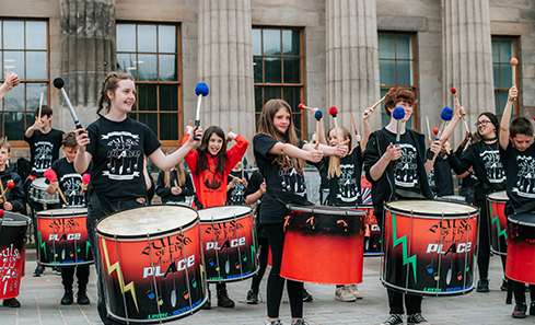 A group of children drumming outside a historic building with pillars at the front