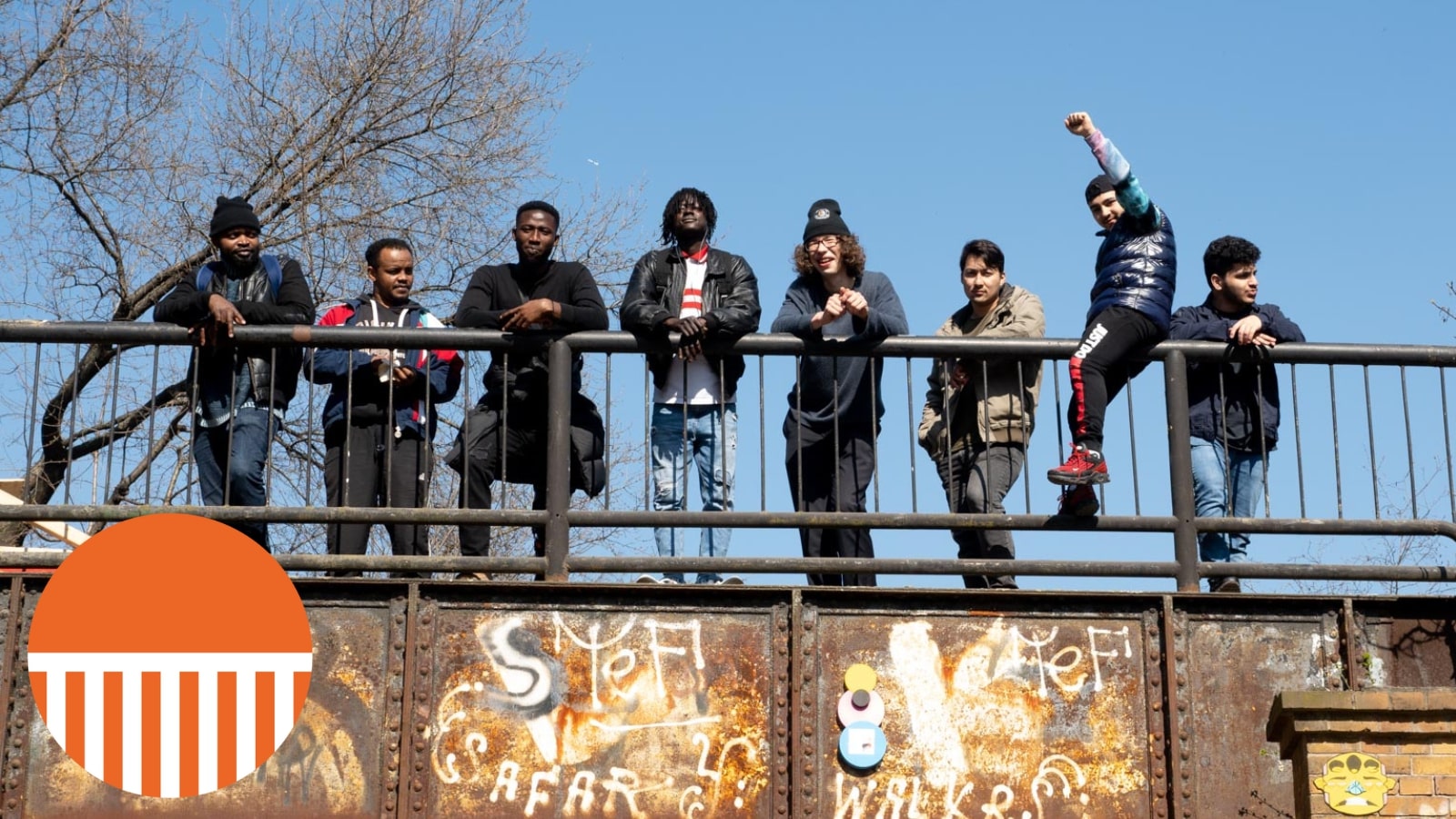 A group of people stand on a bridge looking down over the photographer - they look happy and confident, and one is sitting on the railing of the bridge punching his fist in the air