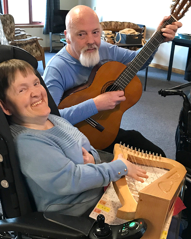 Two people play instruments and grin at the camera - one holding a guitar, the other playing a harp on the tray table of her wheelchair