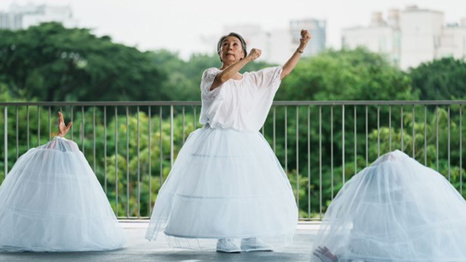 An older woman stands in a white outfit with a full skirt dancing outside next to two other skirts