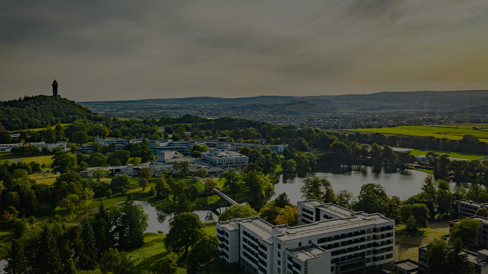 A landscape shot of Stirling which is a very green town with a loch in the centre and a hill nearby where the monument stands - the University is prominent in the forefront of the image, a collection of buildings of varying ages