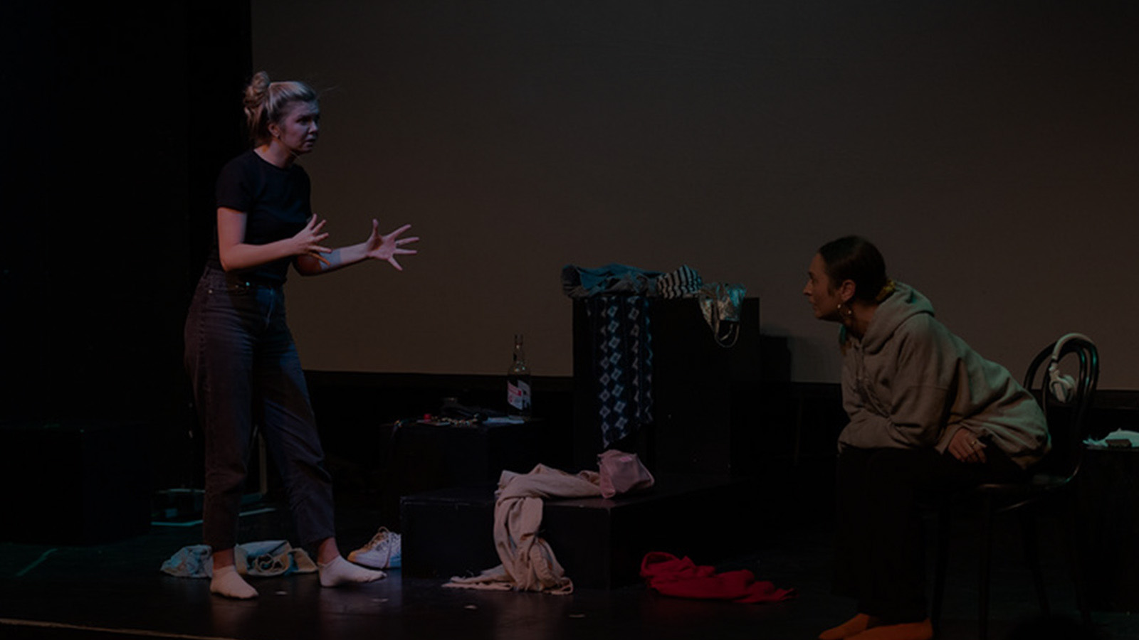 Actors on a stage. One is sitting, one standing. They are surrounded by piles of clothes.