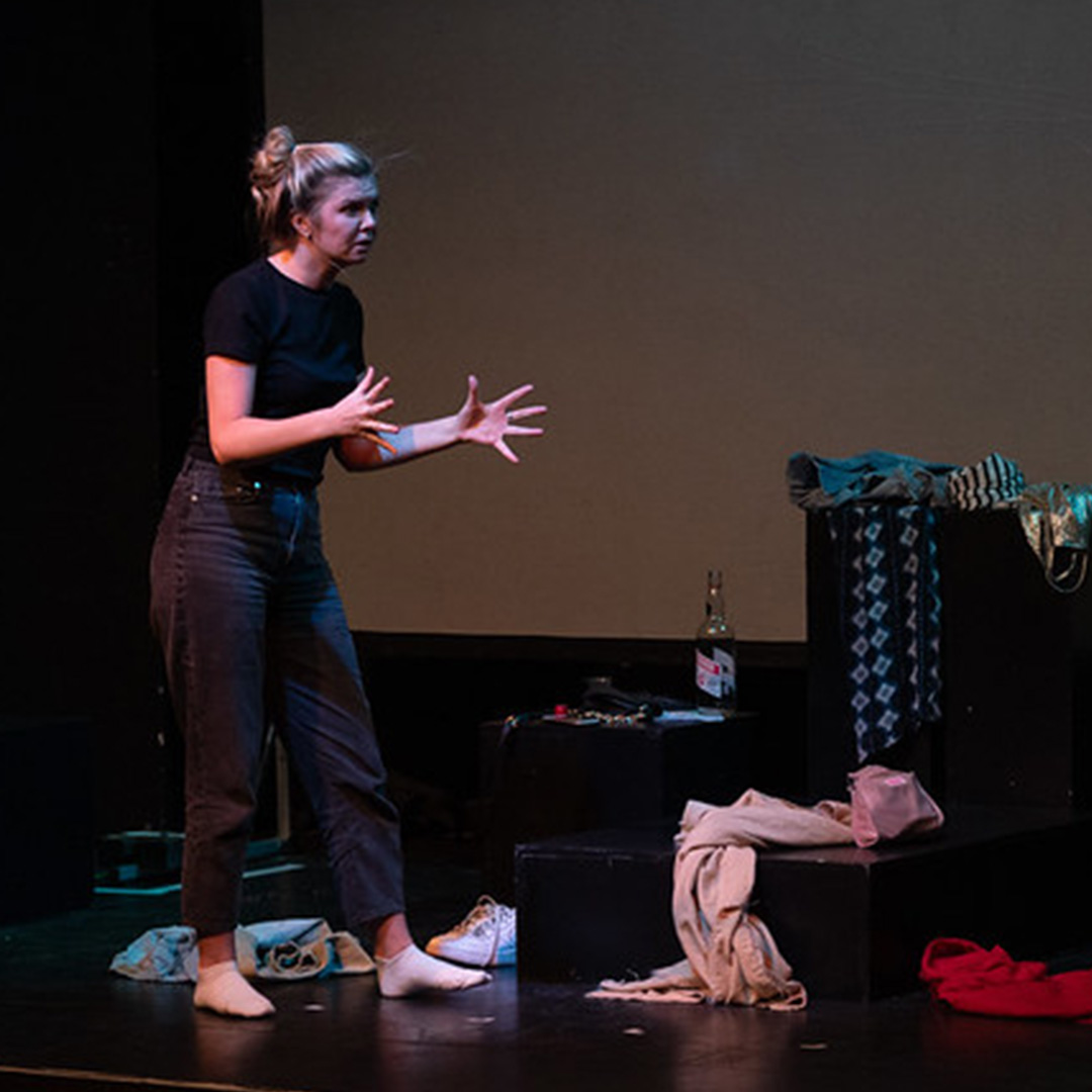 Actors on a stage. One is sitting, one standing. They are surrounded by piles of clothes.
