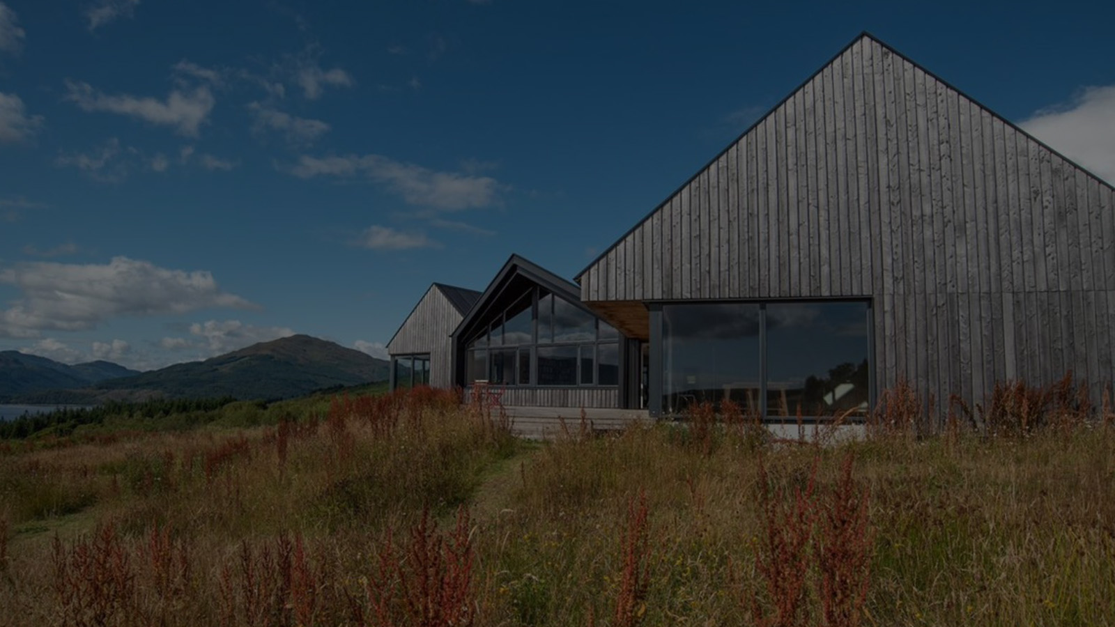 Cove Park. A wooden-clad modern building set in a lush countryside, with a bright blue sky and some fluffy white clouds.