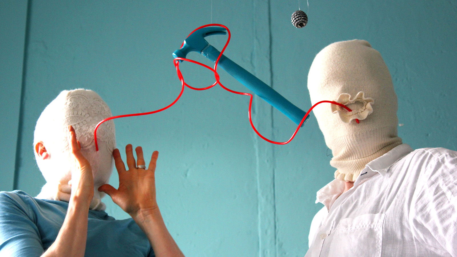 Two people wear white balaclava-like garments on their heads. There are no eye or mouth holes. A red wiggly straw connects from the mouth of one person to the other. A blue hammer is suspended by the straw. A pinecone also hangs from the ceiling.