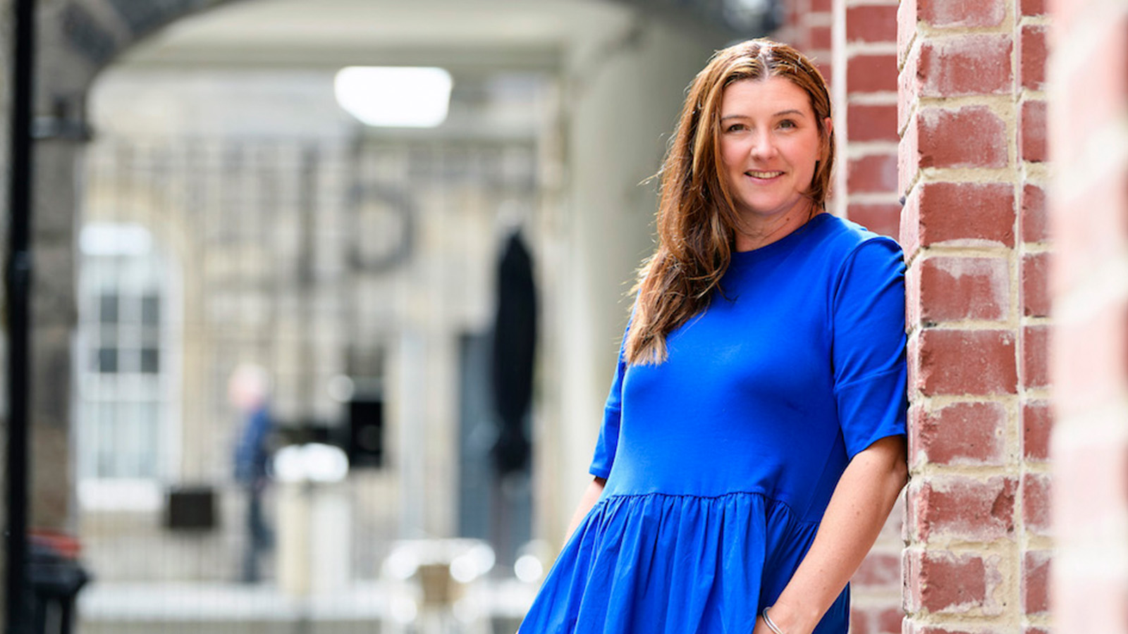 Jenny Niven stands in a bright blue dress leaning against a wall. She smiles gently in the sunshine.