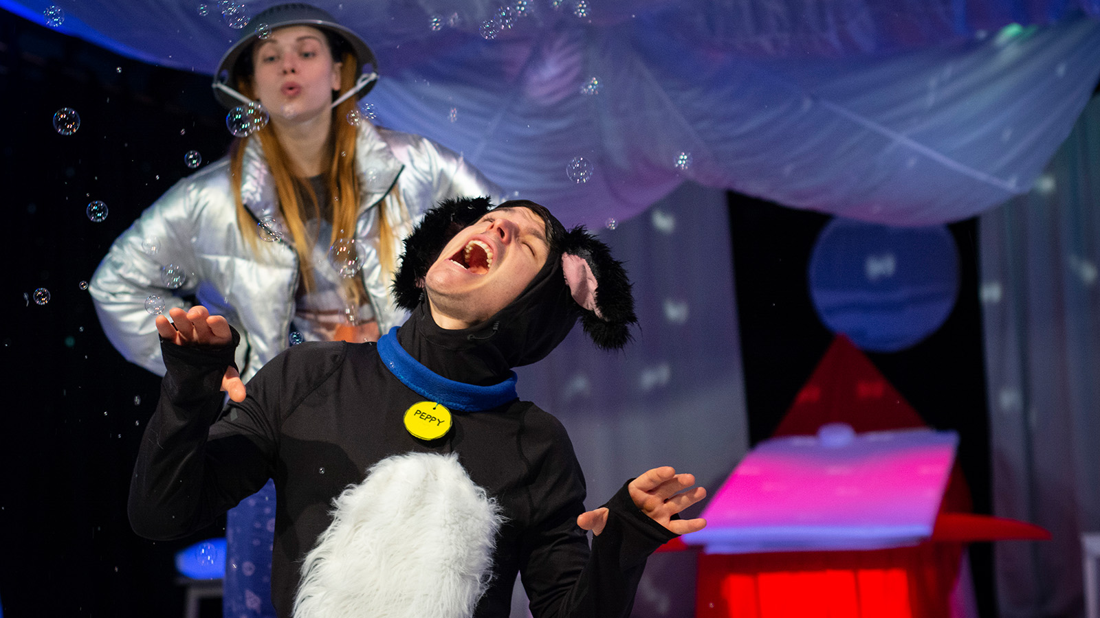 A fun performance where someone dressed as a dog is pretending to sing at the front of the stage, in front of someone else wearing a metal colander as a hat, as though they're dressing up like children, in a colourful environment