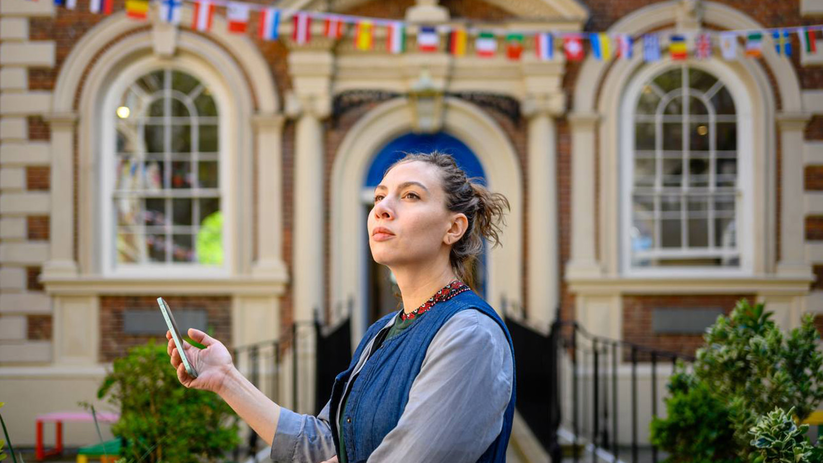 A woman (Veronika Skliarova) holds a smartphone in her hand while looking off into the distance. Behind her is an old brick and stone building with flags of the European nations suspended above the door.
