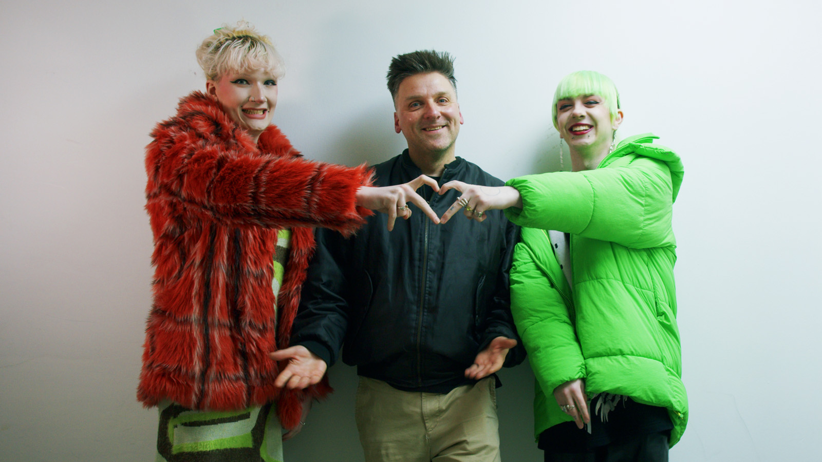 Three people smiling together, two of them have their hands up and are making a heart shape with their fingers