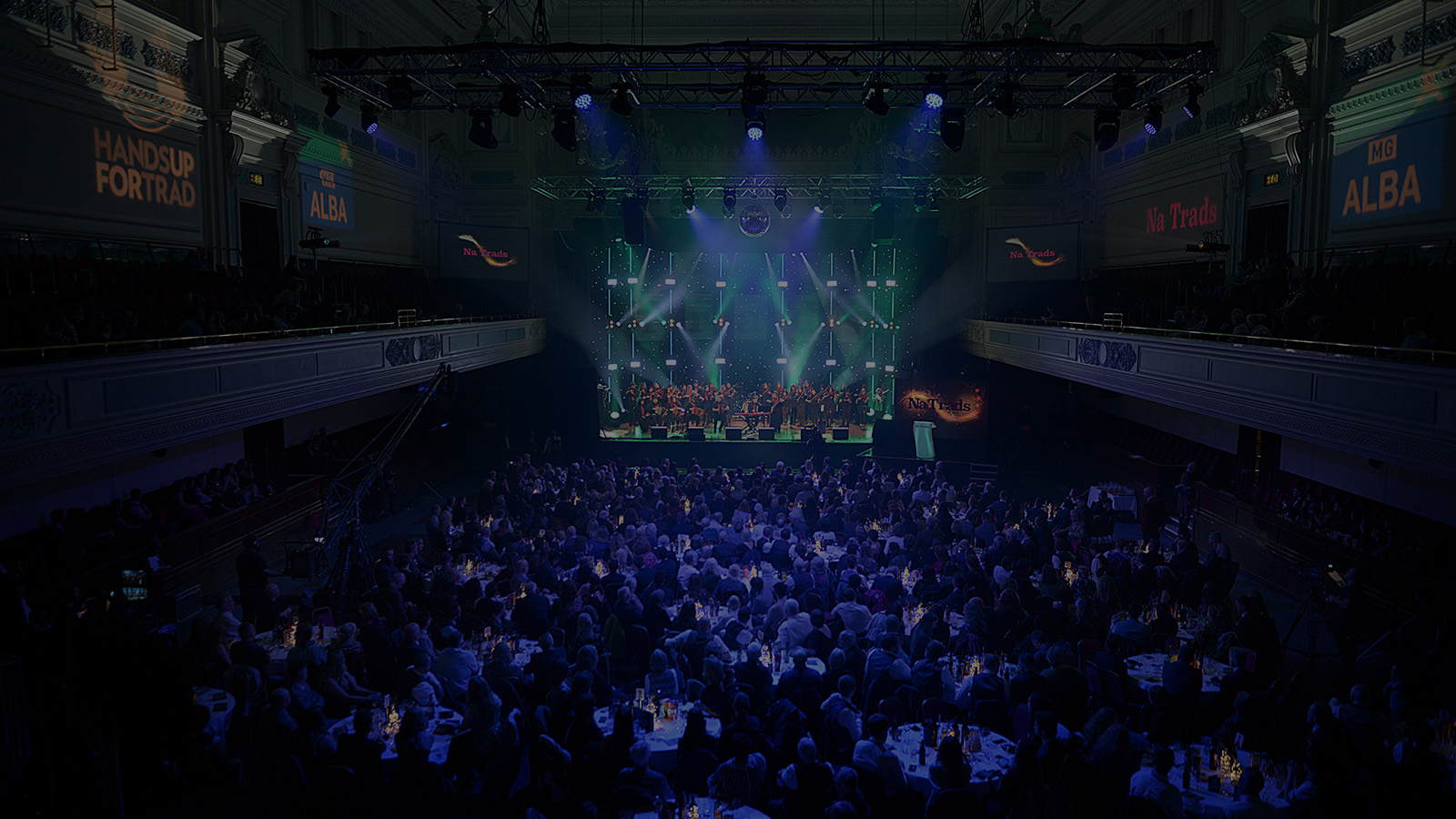 A large audience at an event, a band on stage performing, the auditorium lit in blue and white stage lights