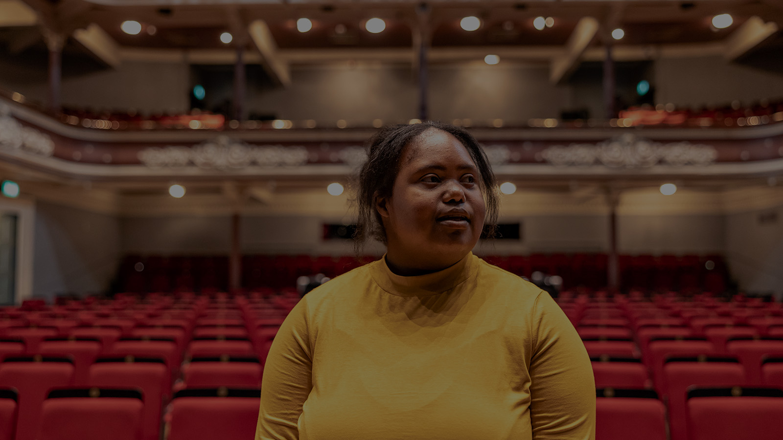 Woman in bright yellow jumper stands in the stalls of a theatre auditorium surrounded by red seats