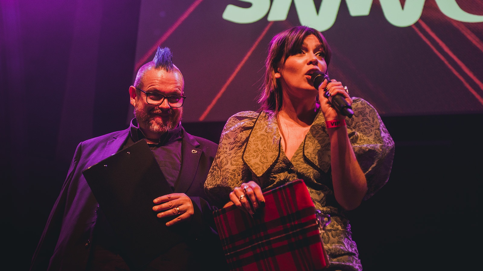 Jim Gellatly and Leyla Josephine as hosts of the 2022 SAMA ceremony stand on stage in front of a large sign that says SAMA holding microphones