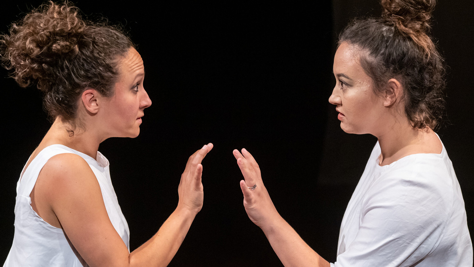 Two women in white t shirts seen in profile, looking at each other as they raise their hands palm to palm