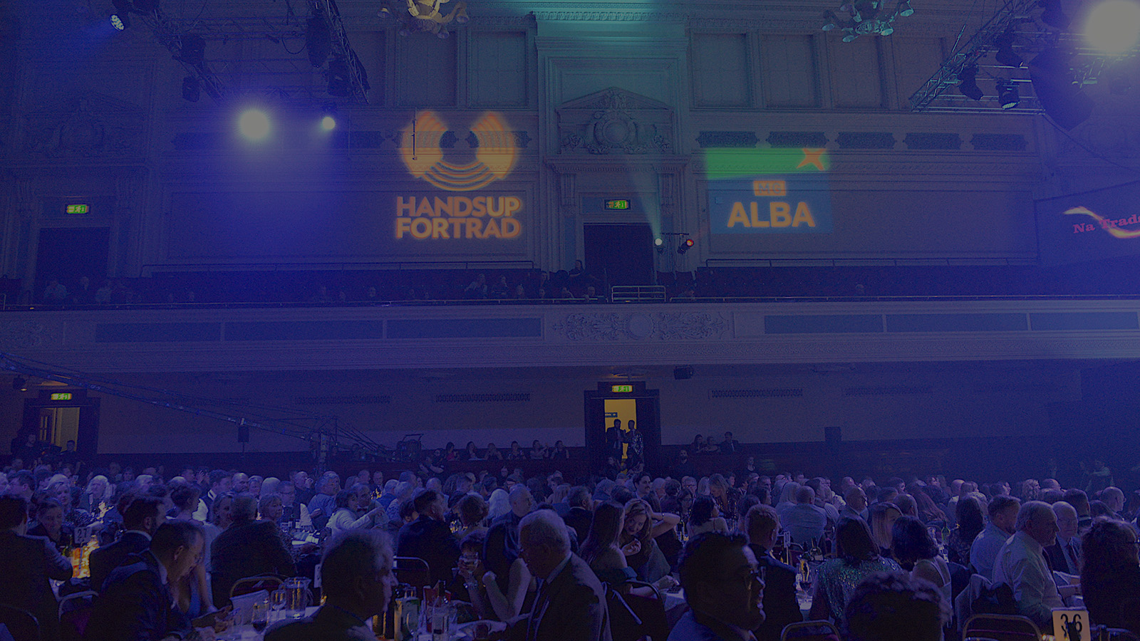 A large audience seated around dinner tables in a atmospheric auditorium, with the logos for the MG Alba Scots Trad Awards projected on to the wall