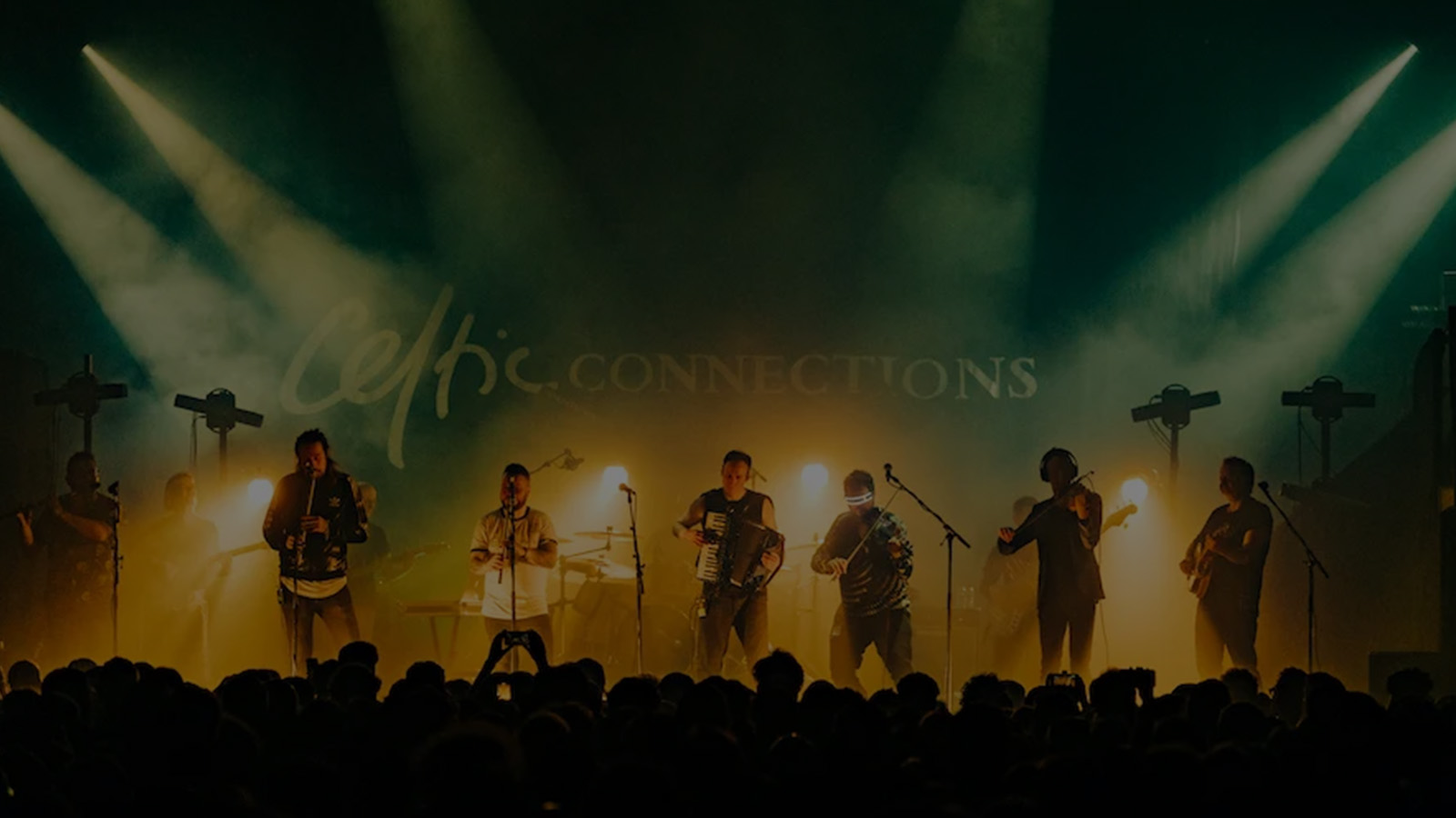 A band perform at Celtic Connections - the image is very atmospheric, with many musicians playing instruments vigorously on stage illuminated with yellow and green lights to a full audience while smoke rises through the air