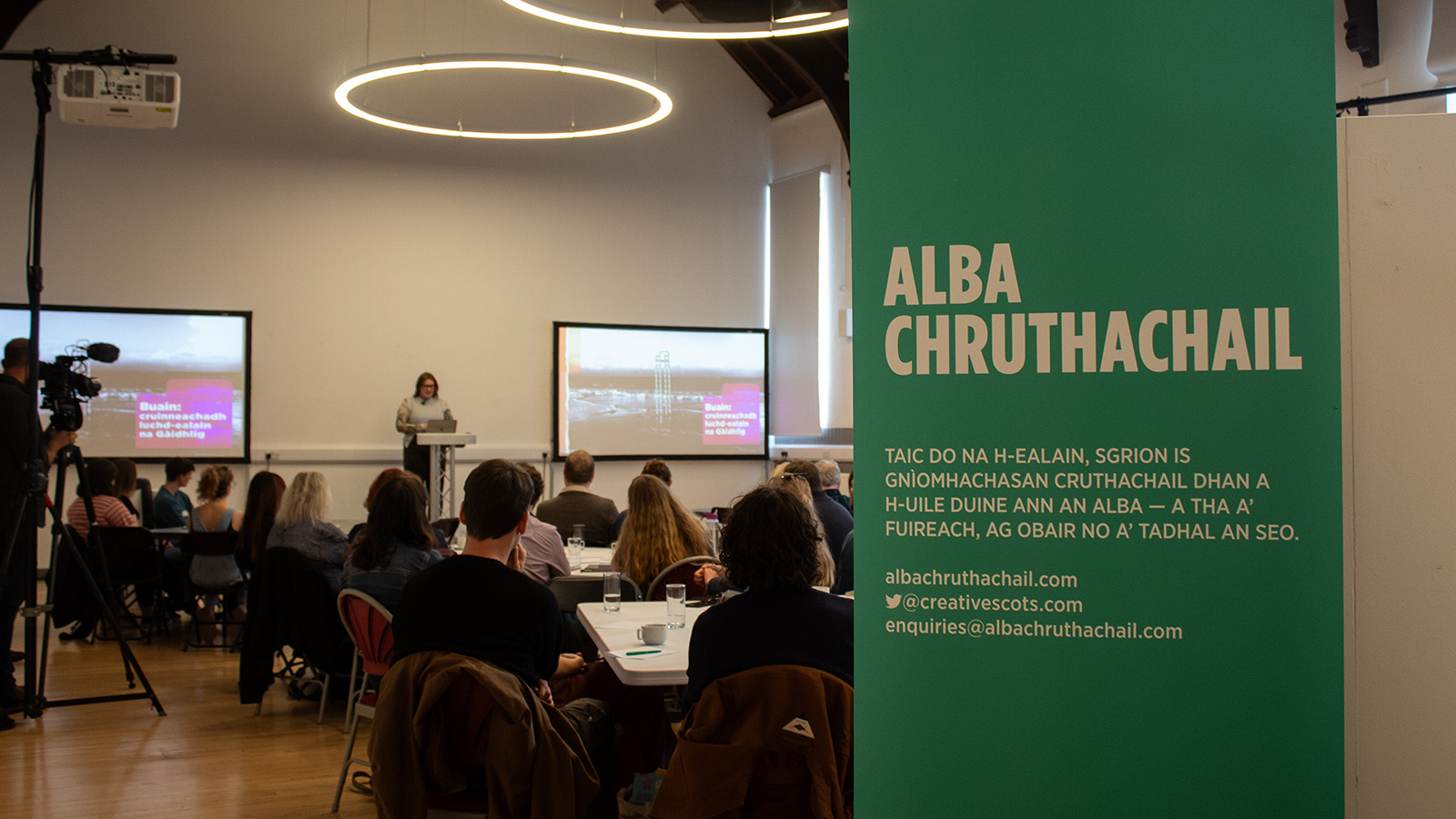 A sign in Gaelic in a room with tables and people speaking at a conference
