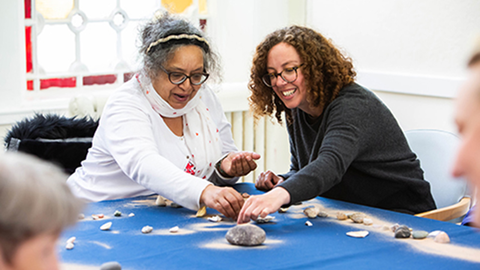 Two women sit at a table smiling while taking part in a creative activity