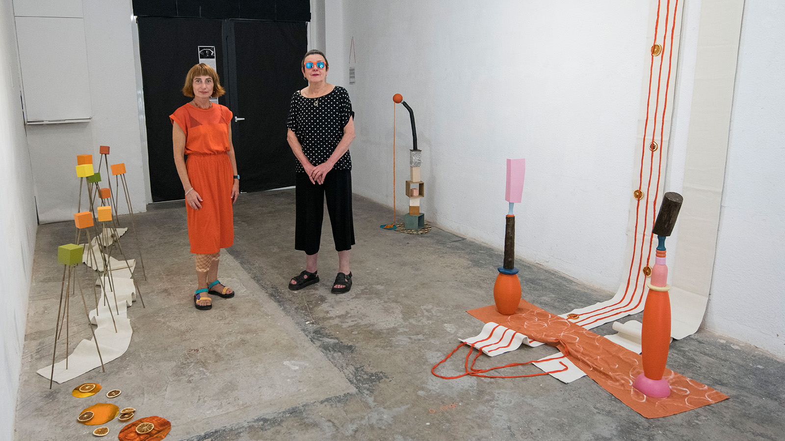Two women, one in a bright orange jumpsuit and one in black, stand in a large white room with various orange and yellow sculptures