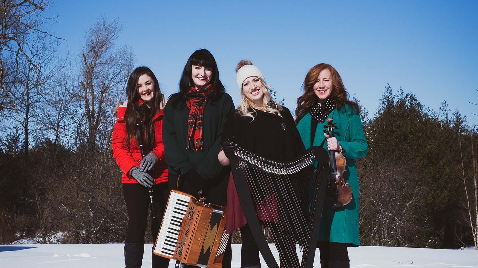 Four women in winter clothing standing in the snow with instruments: a harp, a violin, an oboe and an accordion