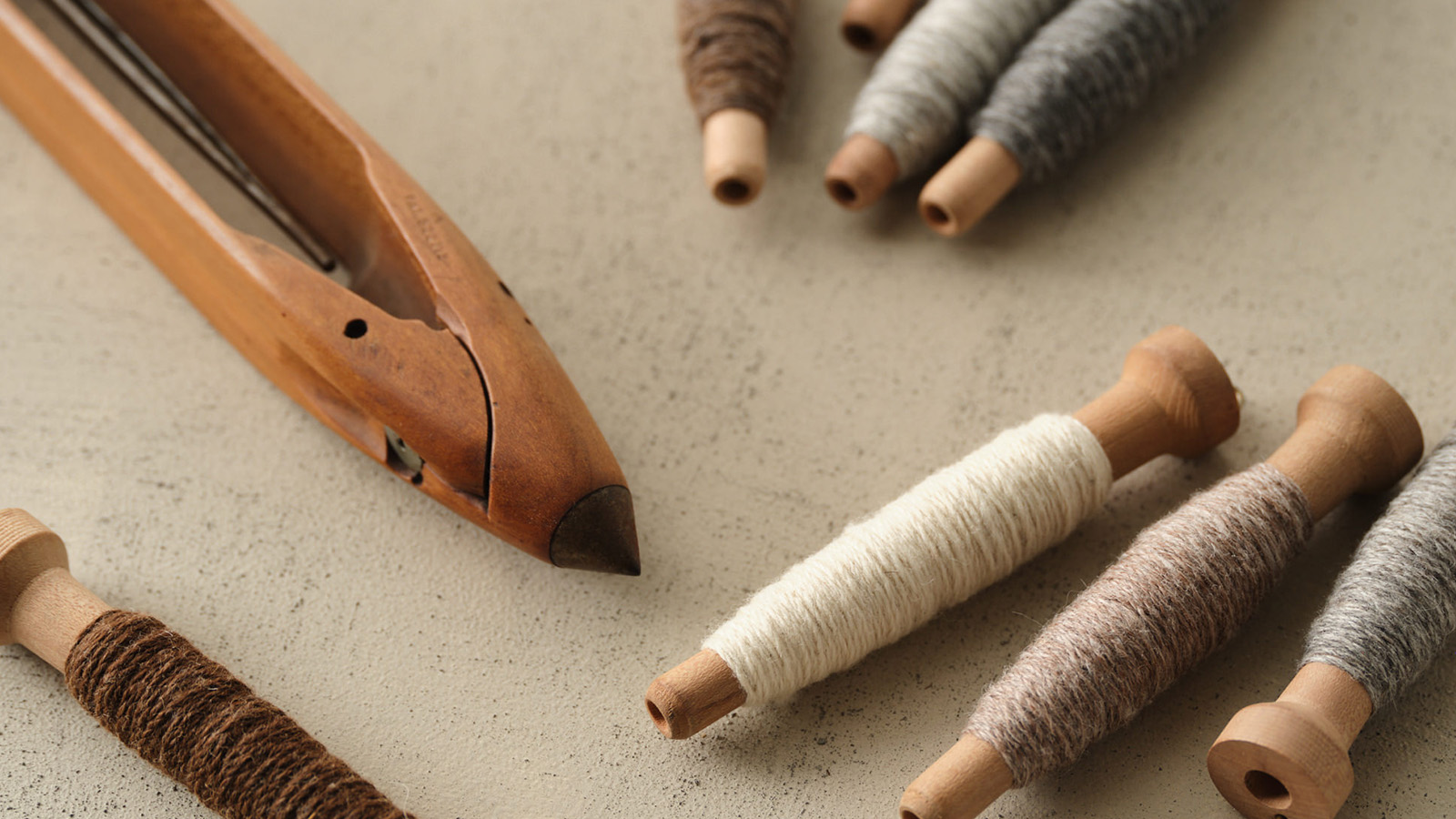 Spools of yarn and tools used in wool craft lay on a table in neutral tones of wood, grey and oatmeal