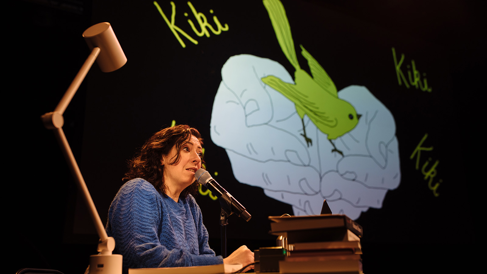 A woman with dark hair speaks into a microphone while she sits at a desk on stage with a spotlight on her. The rest of the stage is in darkness, other than a projection onto the wall behind her to the left, which shows an illustration of a bird in someone's hands