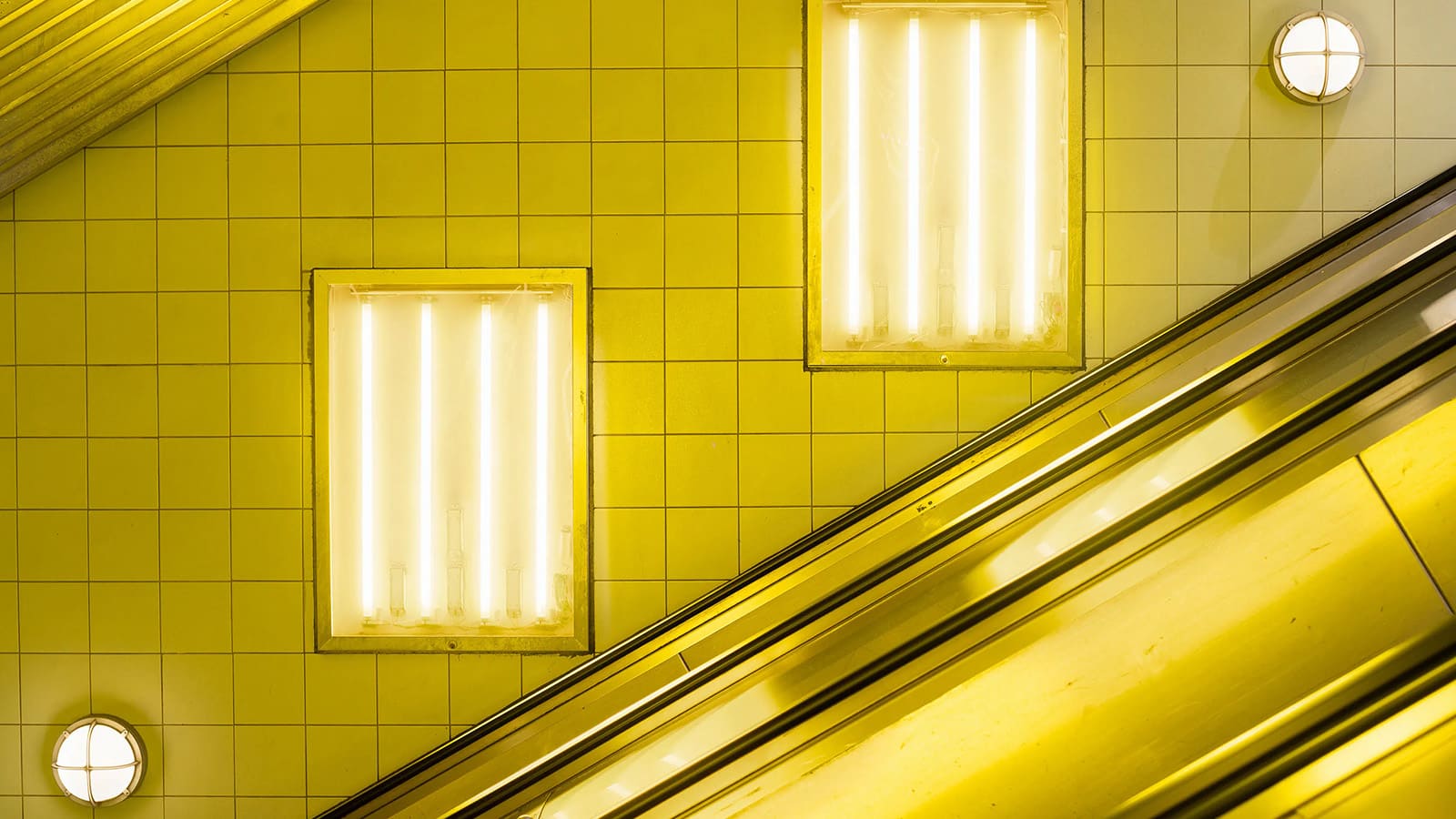 View of an underground or subway escalator, the tiles lit in a bright yellow glow from two strips of neon lights. Thumbnail *