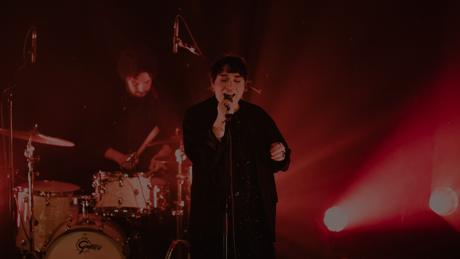 The artist Kitti performs on a stage lit by vibrant red lights. She is holding a microphone and behind her is a person accompanying her on the drums.