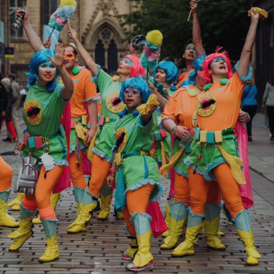 A big group of people wearing colourful superhero costumes and wigs. They're standing in a group posing and showing confidence and strength,on a city street.