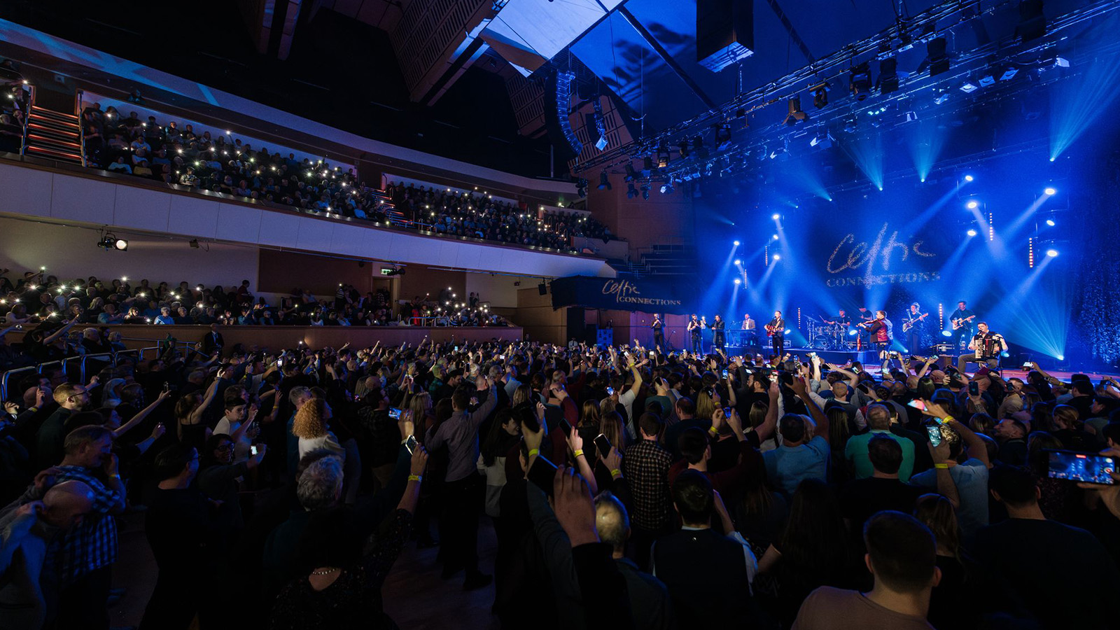 A large crowd of people at a concert, their arms raised, holding phones to capture what's on stage. On the stage, a band of musicians are playing.