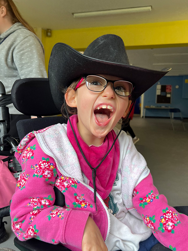 A young girl in a wheelchair wearing glasses and a bright pink kerchief on her neck and a black cowboy hat. She has a big smile on her face.