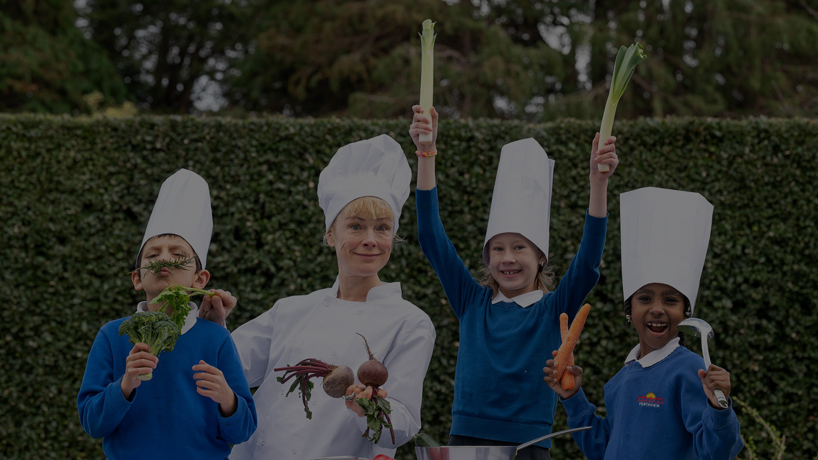 A woman with blonde hair wearing a traditional white chef's outfit and hat and three young children in bright blue jumpers hold up various vegetables