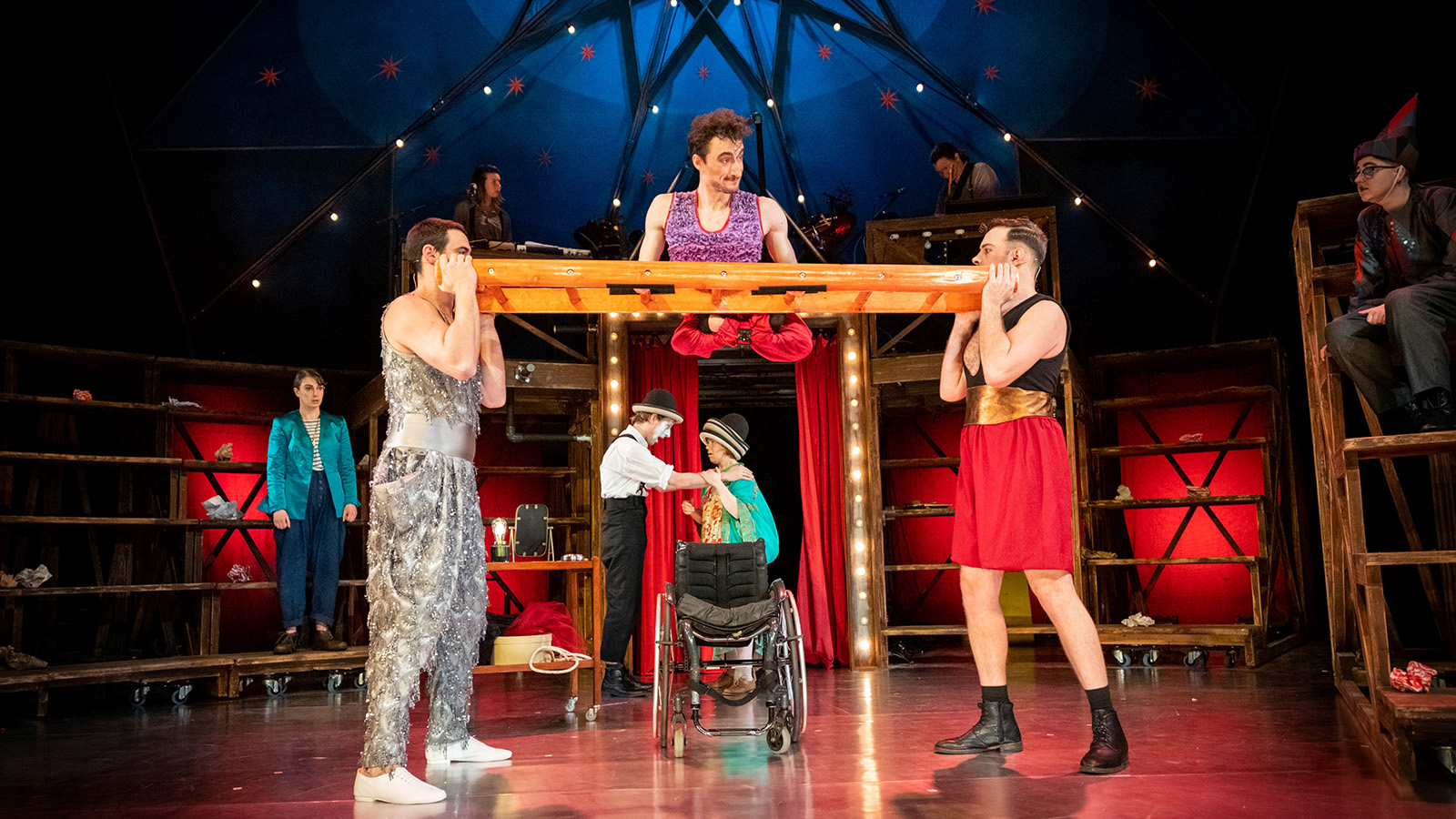 Seven performers in circus costumes. Central performer balances on ladder held horizontally. Empty wheelchair beneath.