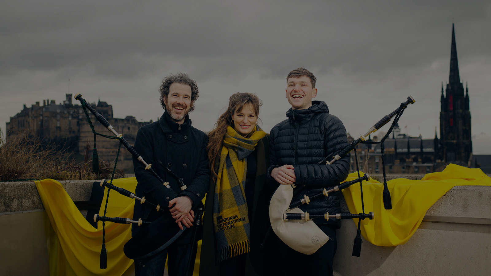 Three people (a young man, an older man, both holding bagpipes,, and a woman) pictured outside with Edinburgh castle in the background. They are musicians Calum MacCrimmon and Conal McDonagh from Breabach and Nicola Benedetti