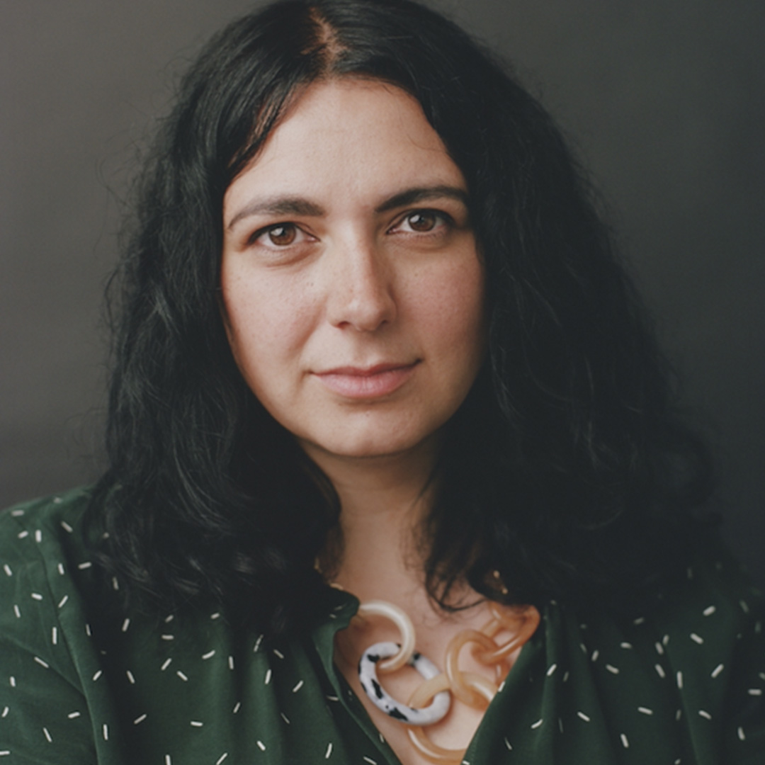 Rachelle Atalla. Rachelle sits looking at the camera in a soft toned portrait. She is wearing a dark green floral top and has long dark curly hair.