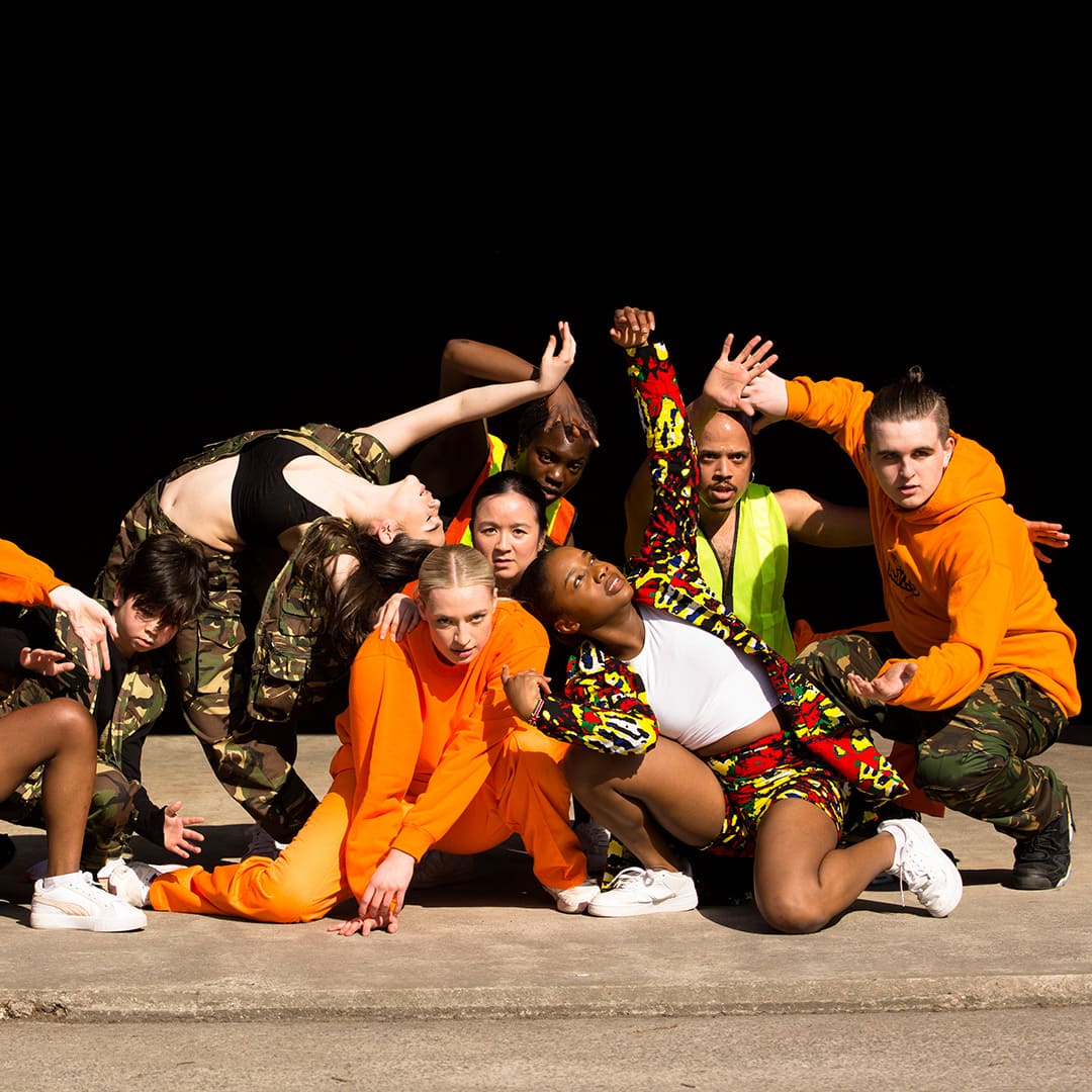 A group of dancers pose on a stage, all dressed in camouflage, or bright orange, green and red