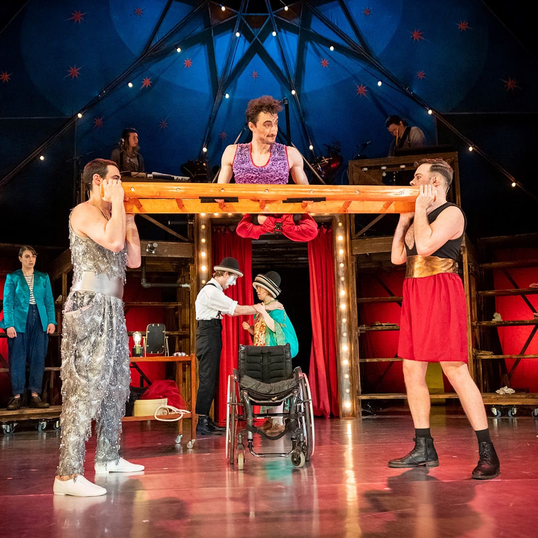 Seven performers in circus costumes. Central performer balances on ladder held horizontally. Empty wheelchair beneath.