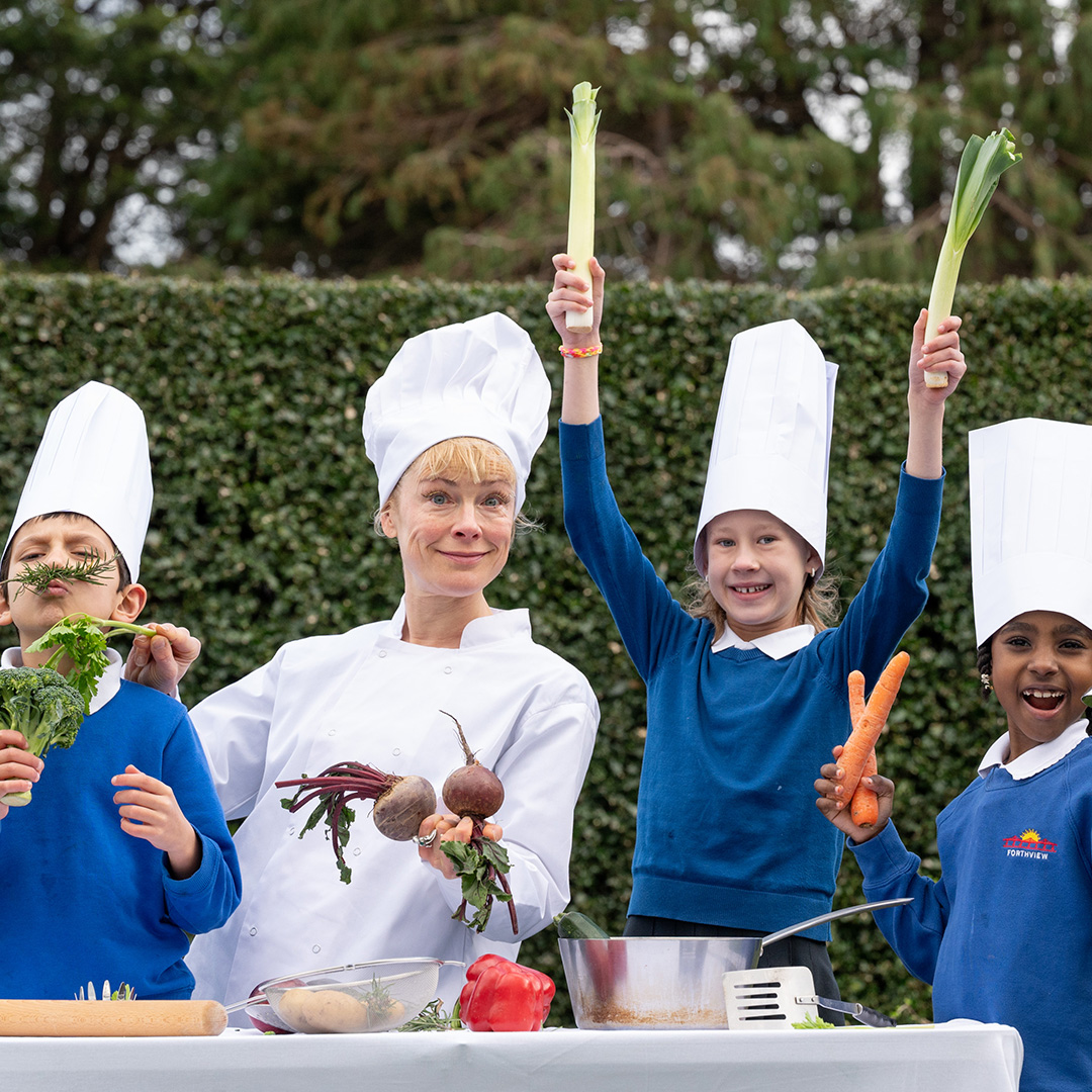 A woman with blonde hair wearing a traditional white chef's outfit and hat and three young children in bright blue jumpers hold up various vegetables