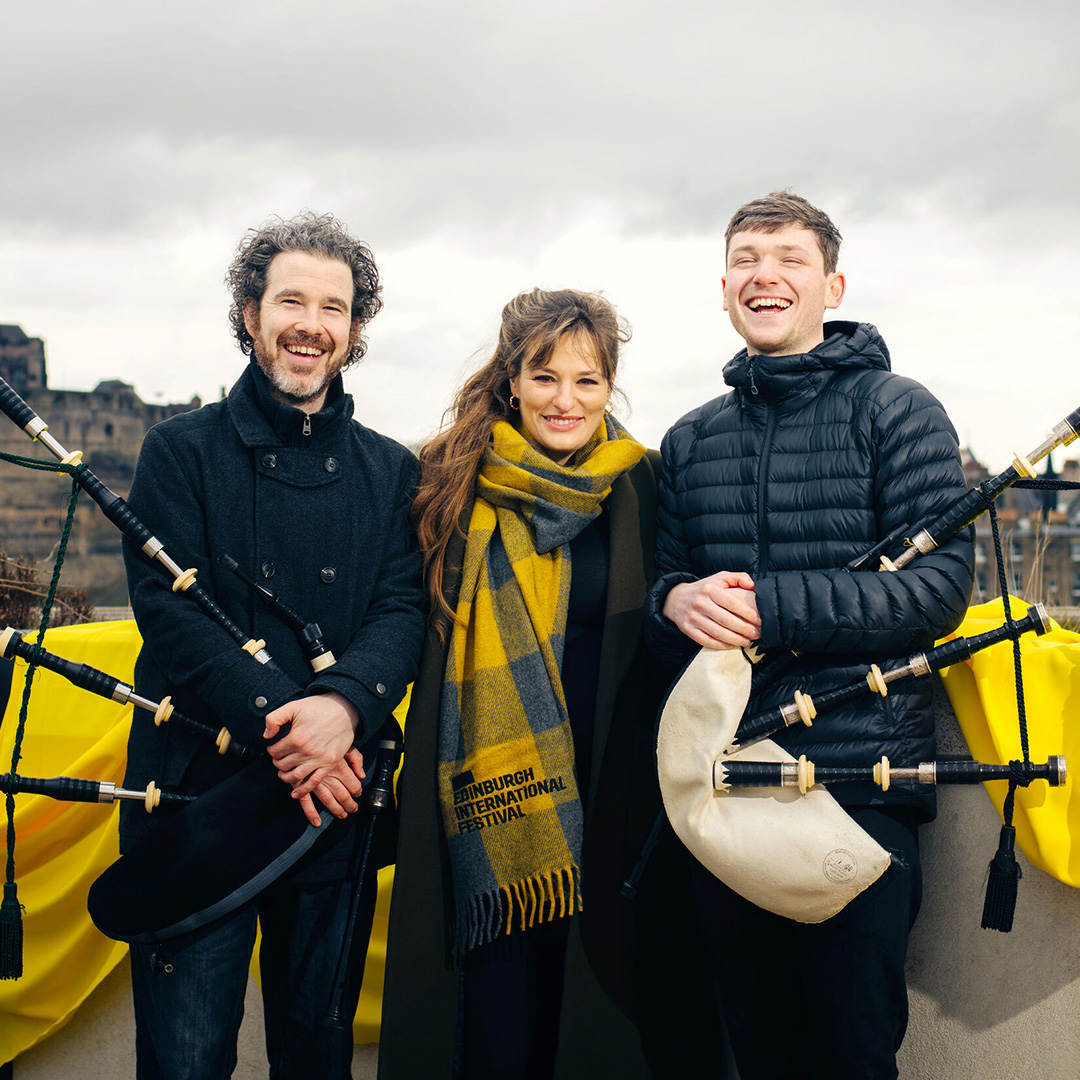 Three people (a young man, an older man, both holding bagpipes,, and a woman) pictured outside with Edinburgh castle in the background. They are musicians Calum MacCrimmon and Conal McDonagh from Breabach and Nicola Benedetti