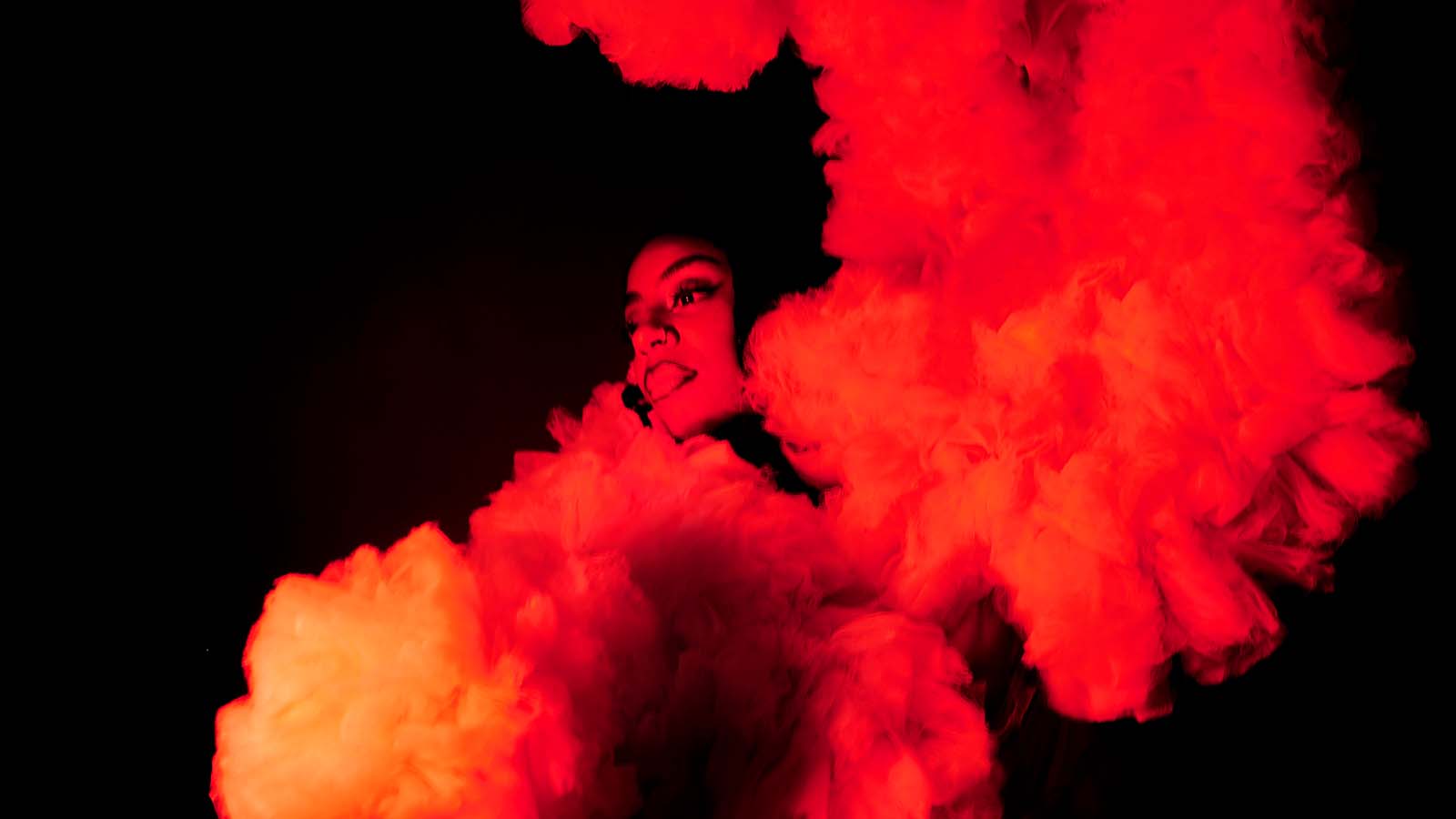 A woman (Male Broomes) surrounded by clouds of what appears to be smoke or cloth, lit in dramatic red and white lighting