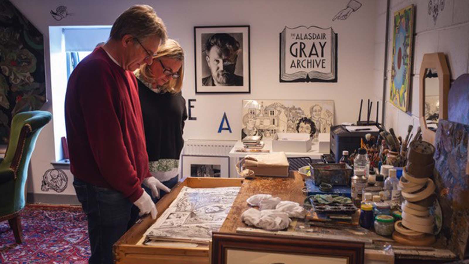 Two people examining the open drawer of an old desk covered in well-used art supplies. On the wall is a sign that reads 'Alasdair Gray Archive.'